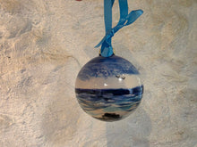 Load image into Gallery viewer, A spherical glass Christmas ornament featuring a painted blue and white seascape with a small bird, hanging from a bright blue ribbon against a textured beige wall.