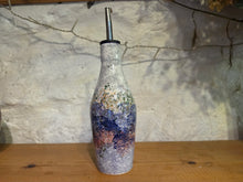 Load image into Gallery viewer, A handmade ceramic bottle with a metallic silver pour spout, featuring a textured, speckled glaze in shades of blue, purple, pink, and white, displayed on a wooden surface against a rustic white plaster wall.