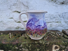 Load image into Gallery viewer, A jug with sky, layers of hills and wild grasses and flowers painted on it. The jug is sat on a piece of sandstone brick with the wall of an old stone building behind it.