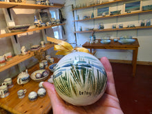 Load image into Gallery viewer, A hand holds a round ceramic ornament in a pottery shop. The ornament is white with green and blue painted maram grass patterns, with the sea in the background, tied with a gold ribbon, and has the word 'Islay' written on it.