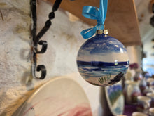 Load image into Gallery viewer, A hand-painted blue and white Christmas ornament featuring a beach scene, hanging from a light blue ribbon on a rustic wooden shelf.