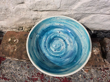 Load image into Gallery viewer, A handmade ceramic bowl with a blue and white spiral glaze pattern, photographed from directly above on rustic wooden blocks against a white-painted brick wall.
