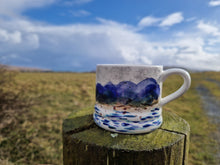 Load image into Gallery viewer, A mug sat on top of a wooden stob. Grass and hills in the background