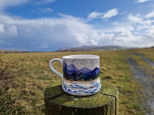Load image into Gallery viewer, A mug on top of a wooden stob with grass and hills in the background