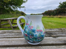 Load image into Gallery viewer, A white ceramic pitcher decorated with a colorful wildflower pattern, resting on a weathered wooden table with a blurred outdoor park background.
