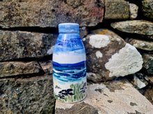 Load image into Gallery viewer, A milk bottle vase painted with blue skies and sea with a rocky, sandy beach. In the background is an old stone wall covered in lichen