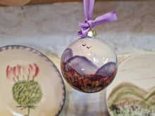 Load image into Gallery viewer, A round bauble with the hills and wild flowers painted on it. A white stone wall in the background with a plate with a pink thistle on it, and another plate with green hills and pink skies painted on it