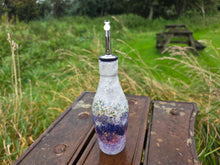Load image into Gallery viewer, A handmade glass bottle with a silver metal pour spout, featuring a white and purple speckled pattern, resting on a weathered wooden picnic table in an outdoor grassy setting.