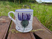 Load image into Gallery viewer, A white ceramic mug with a purple thistle design, resting on a rustic wooden table in an outdoor grassy field.