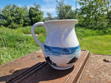 Load image into Gallery viewer, A jug with a painting of the beach with rocks on it and wild grasses, sea and sky. The jug is sitting on a rusty plinth with grass and trees in the background