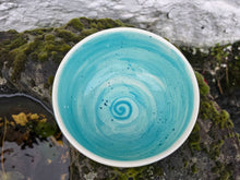 Load image into Gallery viewer, A handmade ceramic bowl with a turquoise blue and white spiral pattern inside, shown resting on mossy rocks near water.
