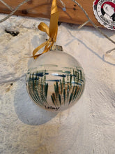 Load image into Gallery viewer, A clear glass Christmas ornament decorated with a hand-painted marram grass and sea scene in green and brown, hanging from a gold ribbon against a textured white wall.
