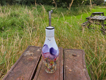 Load image into Gallery viewer, A decorative glass oil bottle with a metal pour spout, featuring a purple and multicolor floral design, set on a rustic wooden table in an outdoor grassy field.
