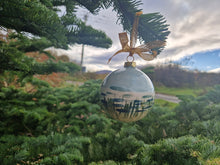 Load image into Gallery viewer, A round bauble with tidal waves, blue sky and marram grass hanging from a fir tree with clouds and sky in the background 
