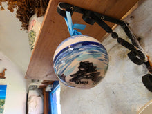 Load image into Gallery viewer, A hand-painted ceramic ornament shaped like a sailboat on water, featuring blue and white waves, and a rocky shoreline, hanging from a black bracket on a wooden shelf in a rustic room.
