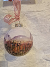 Load image into Gallery viewer, A round bauble with sky, hills and wild flowers hanging against a white stone wall
