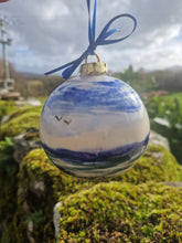 Load image into Gallery viewer, A bauble with deep blue skues, turquoise seas, sand and rocks painted on it set against a moss covered old stone wall.
