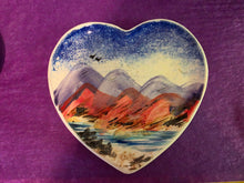 Load image into Gallery viewer, A heart-shaped ceramic dish featuring a hand-painted landscape design with blue mountains, red-orange hills, and a blue sky, set against a textured purple background.
