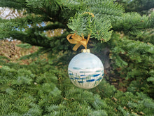 Load image into Gallery viewer, A round bauble with the sea and sand painted on it, along with marram grass. The bauble is hanging with ribbon from a fir tree
