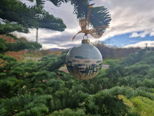 Load image into Gallery viewer, A bauble with the sky sea and marram grass painted on it, set against a sky with blue and cloyds, and a fir tree
