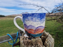 Load image into Gallery viewer, A large ceramic mug with hand-painted mountainous landscape design, predominantly in blue and orange colors, placed on a wooden surface with a field and hills in the background.
