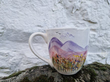 Load image into Gallery viewer, A large mug with hills, grasses and wildflowers painted on it, on a rough piece of wood. A rough stone wall in the background 
