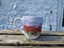 Load image into Gallery viewer, Large mug with a dedign depicting hills and wild flowers on a wooden bench with a white washed wall in the background 
