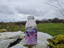 Load image into Gallery viewer, A ceramic milk bottle on a stone wall with grass, hills and sky in the distance 
