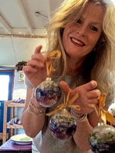Load image into Gallery viewer, A smiling blonde woman holds up three hand-painted glass Christmas ornaments with a marbled purple, blue, and white pattern, each tied with a gold ribbon.
