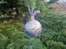 Load image into Gallery viewer, A bauble with sponged pinks and purples hanging with a ribbon with fir tree branches in the background 
