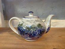 Load image into Gallery viewer, A handmade ceramic teapot with a blue and green floral glaze pattern on a white base, sitting on a wooden surface against a textured gray wall.
