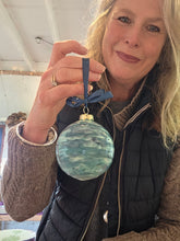 Load image into Gallery viewer, A large round bauble with an abstract turquoise and blue pattern. With splashes of white. The bauble is hanging from a robnon held by a lady in a jumper and waistcoat.
