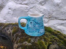 Load image into Gallery viewer, A handmade ceramic pitcher with a turquoise blue and white marbled glaze and dark blue speckles, resting on a mossy rock against a white stucco wall.
