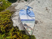 Load image into Gallery viewer, A flat heart decoration with the sky and sea painted on it. A ribbon tied in a bow for hanging it, sat on a rock beside some moss
