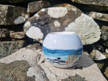 Load image into Gallery viewer, A round tealight holder with the sea and rocks painted on it. With an old stone wall behind it and a lichen covered syone beneath it.
