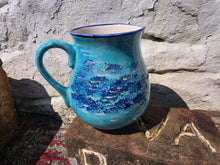 Load image into Gallery viewer, A turquoise blue ceramic pitcher with a blue patterned design on the front, resting on a rustic stone surface against a white brick wall.
