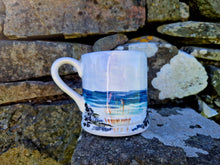 Load image into Gallery viewer, A mug with the sea and rocks painted on it. The mug is sat on a rock with a stone wall covered in lichen in the background 
