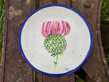 Load image into Gallery viewer, A round white ceramic plate with a blue rim, featuring a painted pink and green thistle flower in the center, resting on a rustic weathered metal surface.
